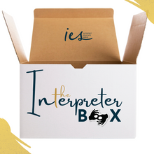 Load image into Gallery viewer, White open box. Front of box has wording "The Interpreter Box". The "o" in Box is replaced with the interpreter symbol. On open flap of box, the IES Interpret Educate Symbol logo. In the upper right corner and lower left corner, gold accents.